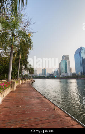 Vista delle palme, la passerella di legno e del lago alla Benjakiti (Benjakitti) Park e i moderni grattacieli di Bangkok, Tailandia. Foto Stock