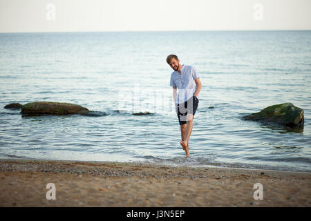Sorridente giovane schizzi di acqua sulla spiaggia. Egli allegramente ride e solleva gli spruzzi di acqua. Foto Stock