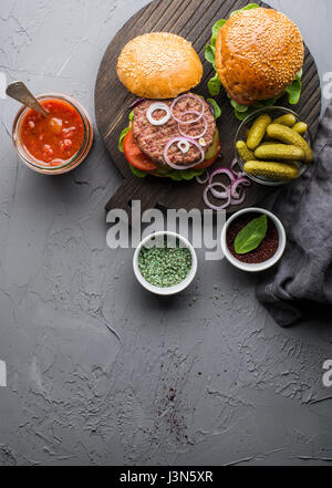 Freschi Fatti in casa gli hamburger sul buio che serve board con il piccante salsa di pomodoro, verde sale, pepe, sottaceti e cipolla su calcestruzzo sfondo a trama. Foto Stock