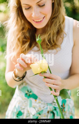 Persone e composizioni floreali concetto - una giovane donna nel giardino estivo fresco di contenimento rosa gialla, dita raddrizza delicatamente i petali del fiore delicato, vestito con una camicia bianca e colorata gonna. Foto Stock