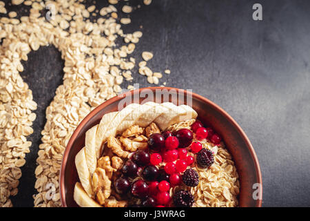 Farina di avena in argilla marrone ciotola con bacche (ribes nero, ribes, more), banane e noci pronto a mangiare, silhouette del cucchiaio su sparse Foto Stock