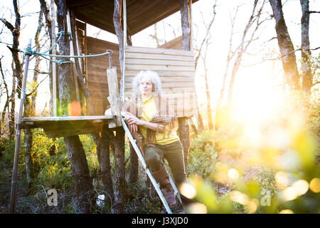 Donna matura appoggiata contro woodland treehouse scaletta al tramonto Foto Stock