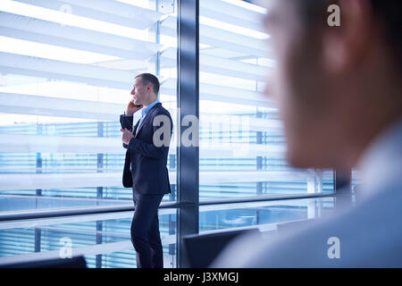 Sulla spalla vista dell uomo d affari rendendo chiamata allo smartphone e guardando attraverso la finestra di ufficio Foto Stock