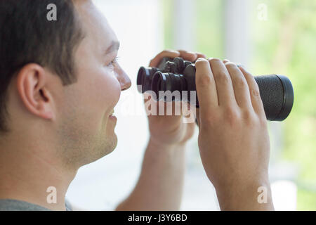 Giovane uomo che sta guardando attraverso una finestra di vetro con un binocolo come si guarda qualcosa di distanza Foto Stock