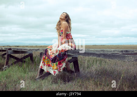 Giovane donna in boho abito maxi seduta sulla sopraelevata passerella in legno in orizzontale Foto Stock