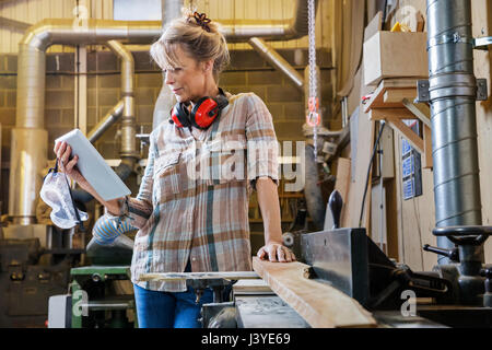 Donna consultando le istruzioni sul tablet mentre utilizzando macchinari in officina in legno Foto Stock