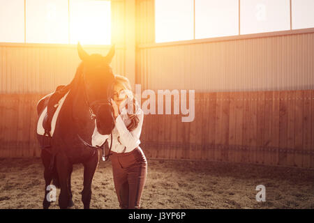 Bellissima ed elegante ragazza bionda sta in piedi vicino al suo cavallo nero medicazione di concorrenza uniforme camicetta bianca maglietta e pantaloni marrone. Piscina portraite in Foto Stock