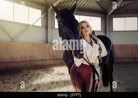 Bellissima ed elegante ragazza bionda sta in piedi vicino al suo cavallo nero medicazione di concorrenza uniforme camicetta bianca maglietta e pantaloni marrone. Piscina portraite in Foto Stock