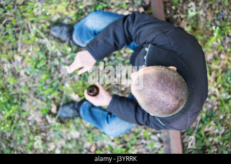 Un uomo seduto con un taglio di capelli corti con una bottiglia e una sigaretta nelle sue mani. Sfocata. Vista da sopra Foto Stock