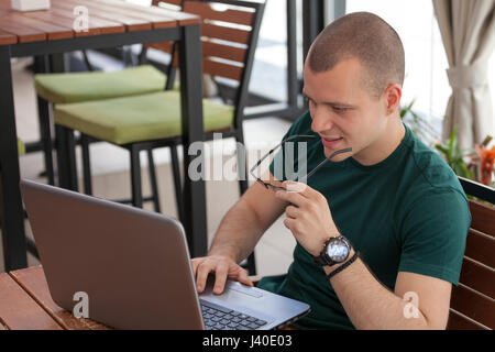 Giovane uomo si siede, tenendo bicchieri in mano e guardando il computer portatile Foto Stock