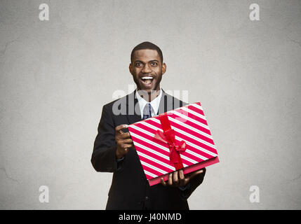 Closeup ritratto giovani felici uomo eccitato apertura confezione regalo rossa, lieti e riconoscenti con ciò che egli ha ricevuto, isolato nero, sfondo grigio. Huma positivo Foto Stock