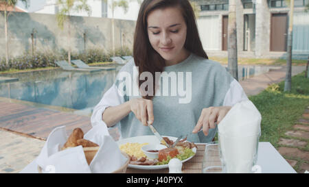 Donna di mangiare la prima colazione con uova strapazzate, salsicce, pancetta, di patata e di pomodoro in grande piastra bianca vicino alla piscina sulla villa di lusso. 4K Foto Stock