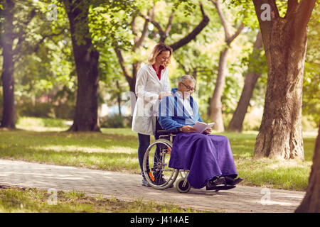 Uomo anziano libro in sedia a rotelle con infermiera sorridente nel parco Foto Stock