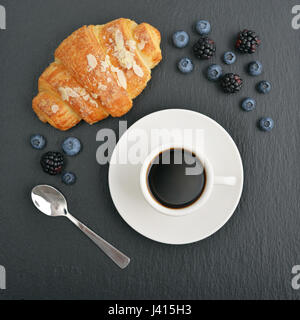 Croissant e caffè con frutti di bosco freschi sul nero ardesia sfondo, vista dall'alto Foto Stock