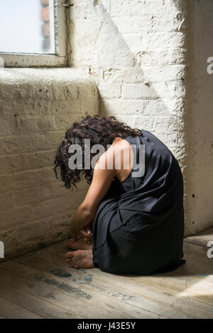 Vista posteriore di una misera giovane donna seduta sul pavimento mediante la finestra Foto Stock