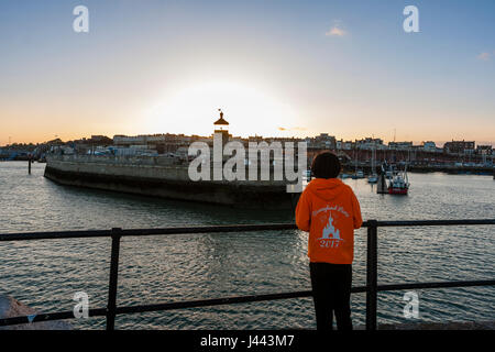 Ramsgate Royal Harbour. Faro alla fine del molo con barche ormeggiate nel porto e dietro che Ramsgate città sul lungomare con il sole che tramonta sulla. Cielo blu chiaro altri poi coppia di piccole nuvole bianche. Il primo piano è un ragazzo adolescente, 14-15 anni, appoggiata contro la ringhiera a guardare il tramonto Foto Stock