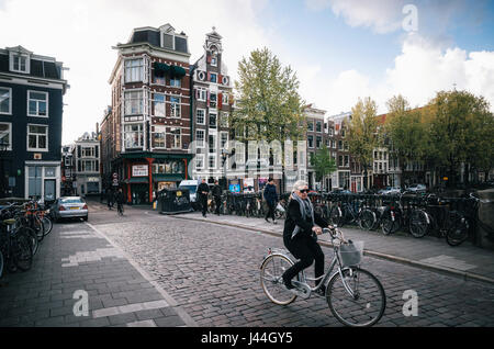 Amsterdam, Paesi Bassi - 25 April, 2017: donna adulta corse in bicicletta in parte storica di Amsterdam con tipiche case tradizionali. Foto Stock