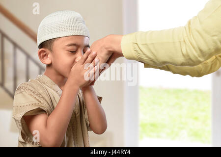 Poco asiatici ragazzo musulmano con abito tradizionale baciare i genitori mano nella casa Foto Stock