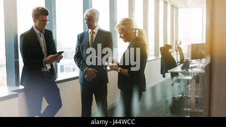 Tre persone in piedi insieme in ufficio con il telefono cellulare e la tavoletta digitale. Professionista aziendale avente una riunione informale nella moderna offic Foto Stock