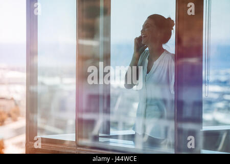 Sorridente imprenditrice matura in piedi all'interno di edificio per uffici e parlando al cellulare. Executive femmina in piedi dal vetro della finestra utilizzando il telefono cellulare. Foto Stock