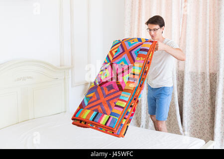 Intimità domestica, homecraft, patchwork, trapuntatura, design concept - uomo bello in blu cielo shorts e bicchieri rende il letto con brillante coltre multicolore realizzato in diverse patch Foto Stock