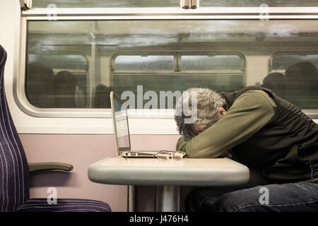 Treno UK. Uomo maturo con il computer portatile a dormire la sede della finestra sul treno vuoto con la pioggia sulla finestra. In Inghilterra. Regno Unito Foto Stock