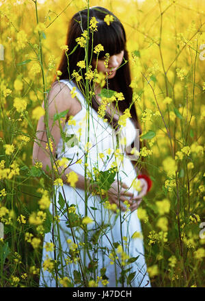 Bella incinta giovane donna asiatica con lunghi capelli neri che indossa un abito bianco mentre tiene il suo ventre come essa si erge in un campo di giallo senape fiori un Foto Stock