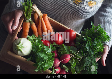 Primo piano della giovane donna caucasica con tessuto grigio maglione tenendo una grande cassa di legno pieno di materie appena raccolto di ortaggi Foto Stock