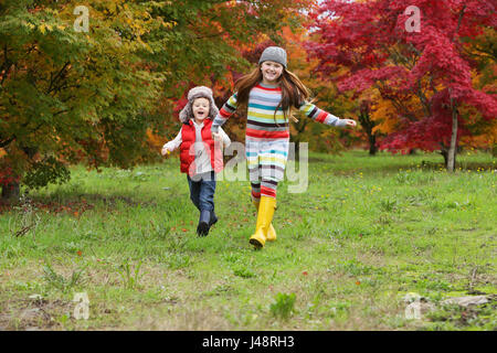 Un ragazzo giovane e ragazza che indossa stivali in gomma e vestiti colorati correre attraverso Un campo che tiene le mani con gli alberi in colori luminosi... Foto Stock