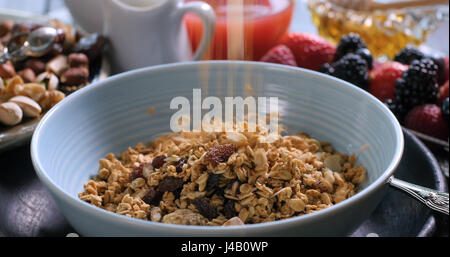 Cereali per la prima colazione che cade su di un recipiente con bacche e frutti secchi sfocati sullo sfondo Foto Stock