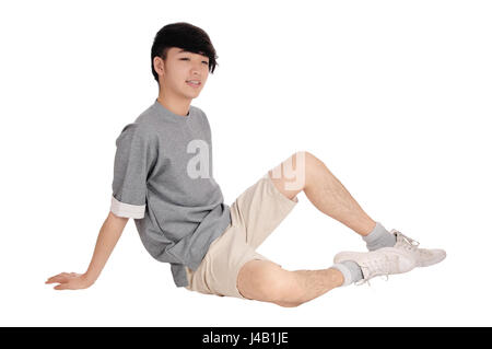 Un bel giovane uomo asiatico seduto in pantaloncini corti e grigio di una t-shirt seduta in studio sul pavimento, isolato per sfondo bianco. Foto Stock