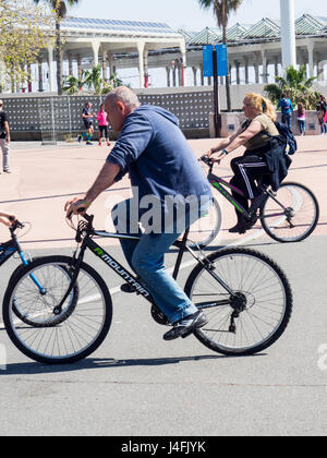 Una coppia di mezza età in bicicletta lungo la spiaggia di Barcellona di fronte a un parco di divertimenti, Barcelona Bosc Urbà. Foto Stock
