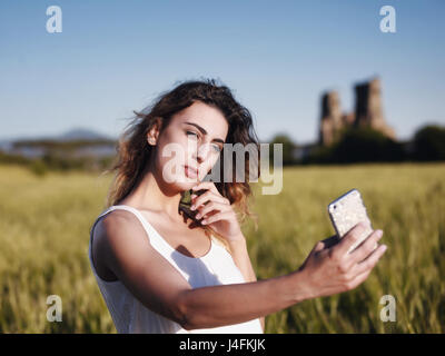 Una giovane ragazza bionda al park, si allunga il suo braccio e con un telefono cellulare lei prende una foto nel paesaggio Foto Stock