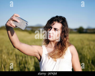 Una giovane ragazza bionda al park, si allunga il suo braccio e con un telefono cellulare lei prende una foto nel paesaggio Foto Stock