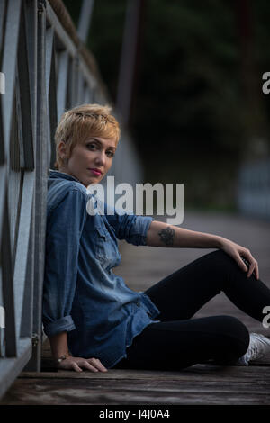 Moderno e alla moda giovane donna con un tatuaggio sul suo braccio seduta rilassante su un legno vecchio e ponte di ferro in abiti casual volgendo lo sguardo pensieroso a t Foto Stock