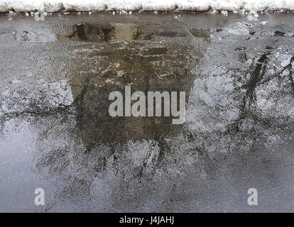 La pozzanghera con la riflessione di una grande casa di mattoni su asfalto, alberi accanto alla casa e il bordo del bianco della neve in inverno. Foto Stock