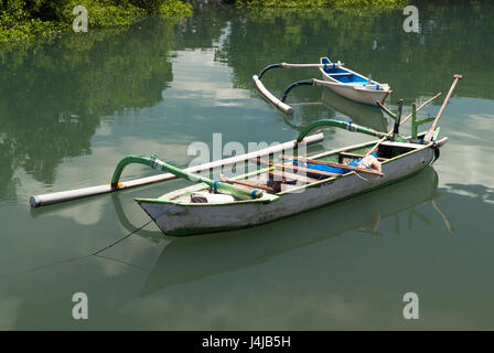 Balinese barca di legno con una unilaterale contrappeso, bianco, legato con una corda alla riva, in background è un altra barca, riflessi nell'acqua Foto Stock