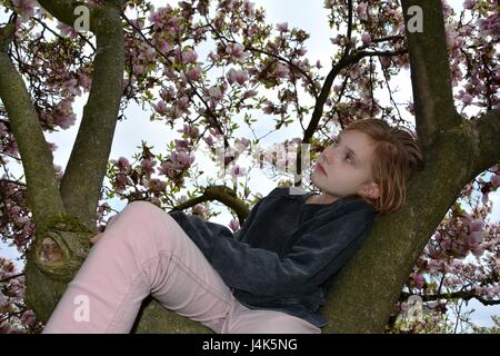 Giovane ragazza si siede in un albero di magnolia e guarda a parte Foto Stock