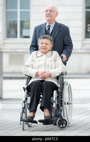 Senior uomo spinge la sua moglie disabili attraverso la città Foto Stock