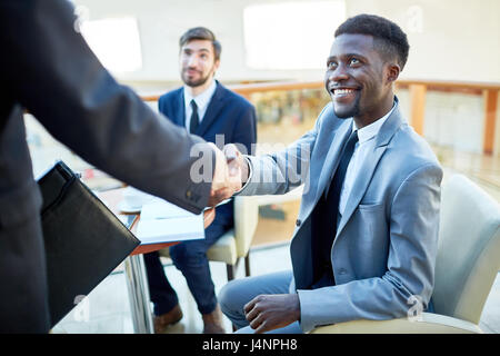 Ritratto di giovane sorridente imprenditore afro-americana si stringono la mano con il partner, seduti al tavolo di riunione Foto Stock