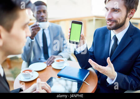 Ritratto di giovane moderno imprenditore con entusiasmo la presentazione mobile app a colleghi mostrando loro smartphone durante la riunione in cafe Foto Stock