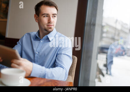 Ritratto di giovane uomo bello sedersi a mensa nel cafe, guardando fuori della finestra e sorridente in daydreaming Foto Stock