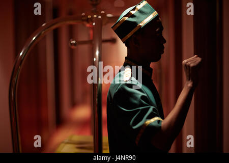 Vista laterale verticale del facchino africani a bussare alla camera di albergo porta consegna bagagli nel corridoio dim Foto Stock