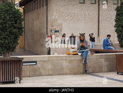 Una ragazza giovane donna seduta su calcestruzzo muro di pietra doppia coppia due coda di cavallo panini esterno guardando giù al telefono mobile square plaza Valencia Spagna Foto Stock