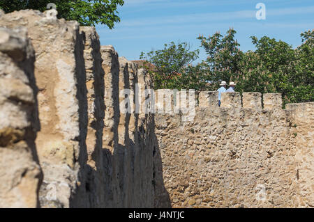 Due uomini turisti persone con cappelli cappello in cima a piedi lungo la parte superiore old antica pietra medievali storiche mura della città dentelli rovine a Segovia, Spagna Foto Stock