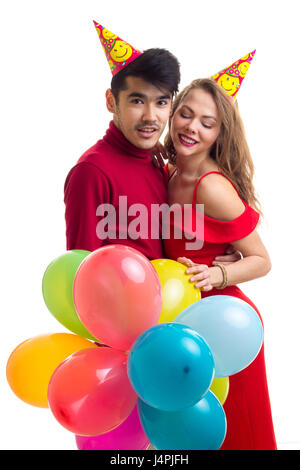 Giovane donna attraente con lunghi capelli castani in abito rosso e il giovane uomo bello in maglietta rossa con celebrando cappelli tenendo molti di palloncini colorati su Foto Stock