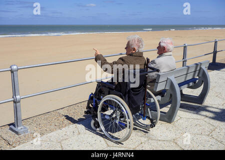 Anziani disabili donna in carrozzella e pensionati marito seduto su una panchina alla passeggiata lungo la costa in una fredda giornata di sole in primavera Foto Stock