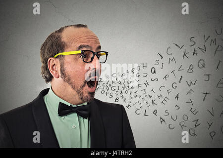 Vista laterale verticale medio di età compresa tra i business man parlando con alfabeto lettere provenienti al di fuori della bocca aperta isolata parete grigio Sfondo. Volto umano expressio Foto Stock