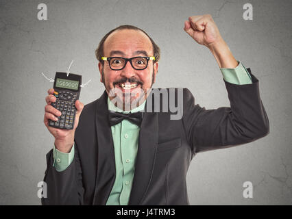 Emozionato happy businessman che mostra la calcolatrice con milioni di segno di numero sullo schermo isolato sul muro grigio office background. Volto umano espressione emozione Foto Stock