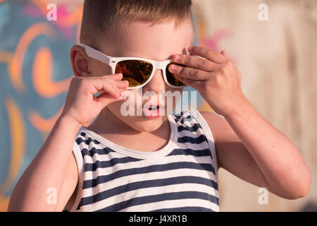 Grazioso piccolo ragazzo con occhiali da sole bianco e sailor stripes vest sul muro di graffiti sullo sfondo di toccare i suoi occhiali da sole Foto Stock